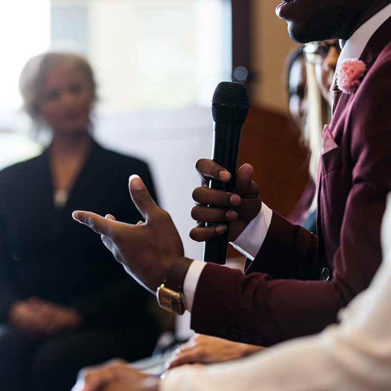 A man holds a microphone in one hand, gesturing with the other, speaking to a group of people. Image is representative of a panel discussion for Natilik's "Spring Clean your Customer Experience" Showcase and Panel Discussion